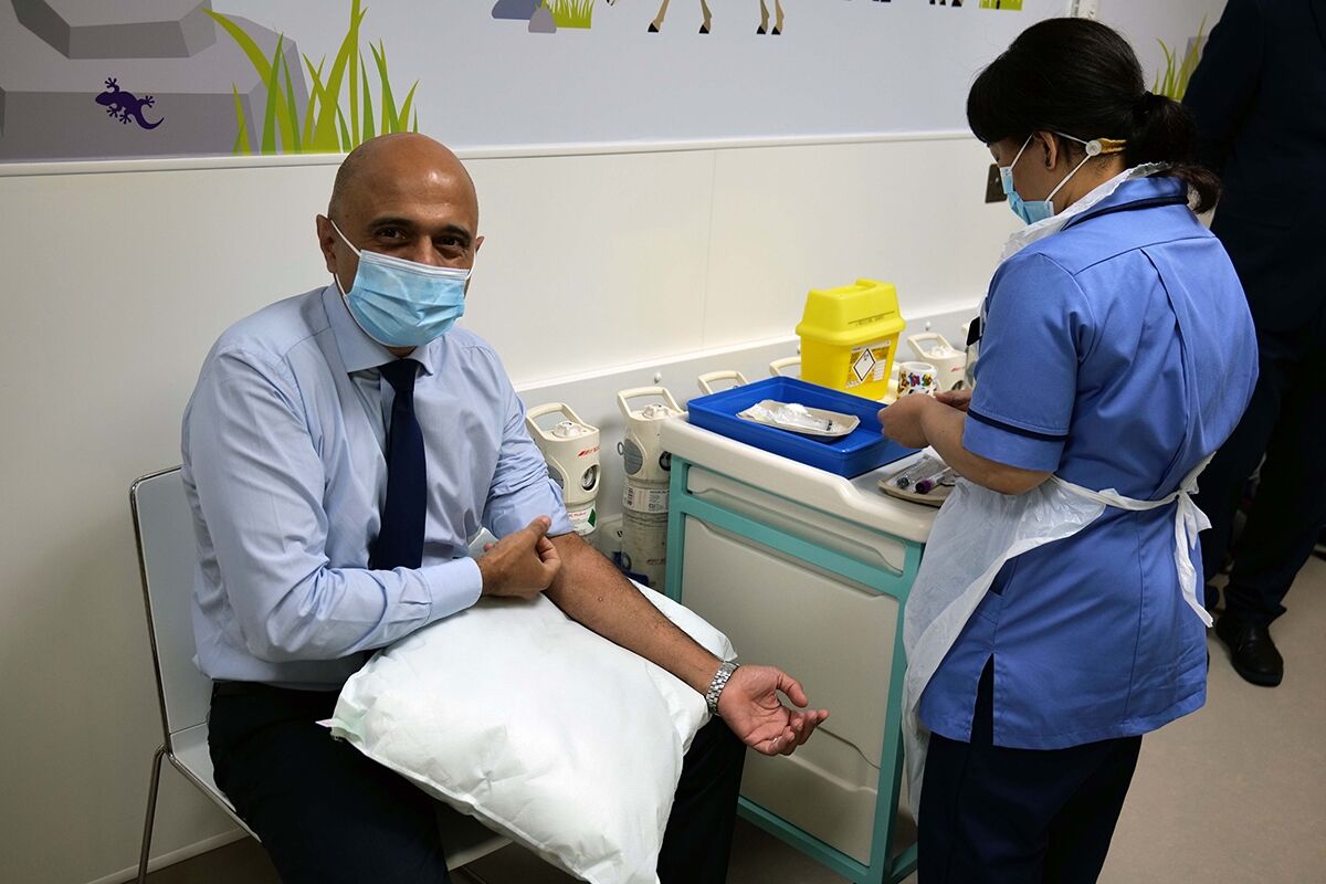 Sajid Javid sitting in a chair facing the camera and rolling up his shirt sleeve whilst a nurse prepares to vaccinate him